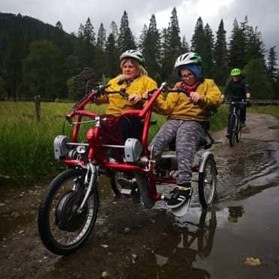 a woman and a young boy riding an adaptive bike together near a forest